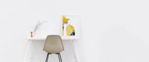 Bild eines Schreibtisches an einer weißen Wand mit einem Stuhl davor. Auf dem Schreibtisch steht ein großer Bilderrahmen mit einer Illustration von einer Frau.