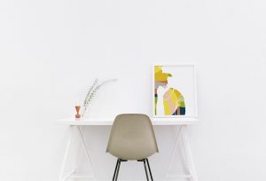 Bild eines Schreibtisches an einer weißen Wand mit einem Stuhl davor. Auf dem Schreibtisch steht ein großer Bilderrahmen mit einer Illustration von einer Frau.